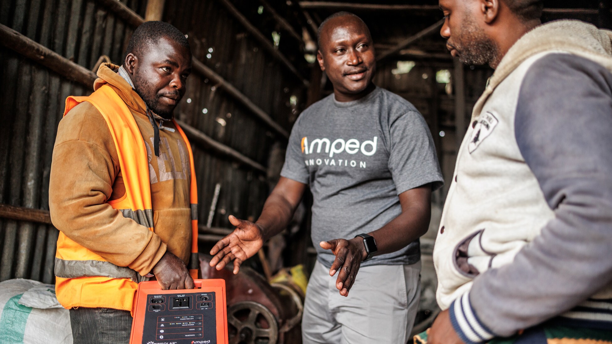 Amped Innovation representative wearing Amped-branded grey t-shirt explains benefits of solar generator to two men standing on either side of him. Man to his right is holding the Amped generator, wearing high-viz yellow vest and orange hoodie; man to his left is wearing a two-toned grey hoodie, looking at the generator while listening.