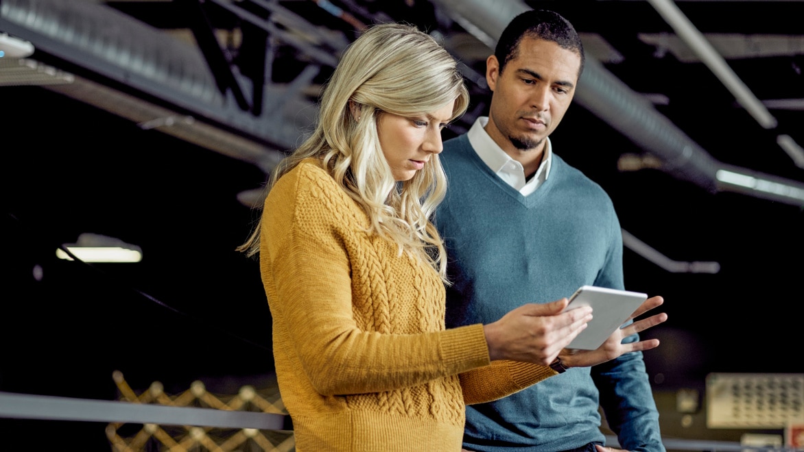 Two colleagues standing together, looking at a tablet and discussing the latest security advisories in an office setting.