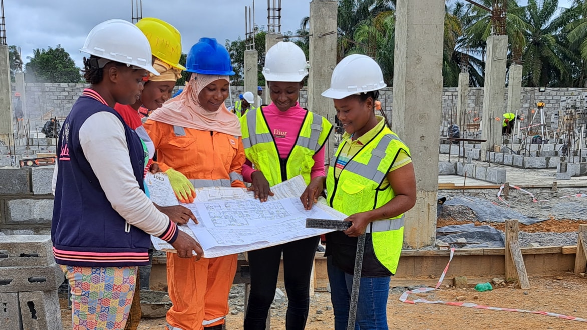 Five BHI women construction workers standing in a semi-circle, holding and examining a large paper schematic and smiling, at the construction site of the new BHI MCOE in Sierra Leone.
