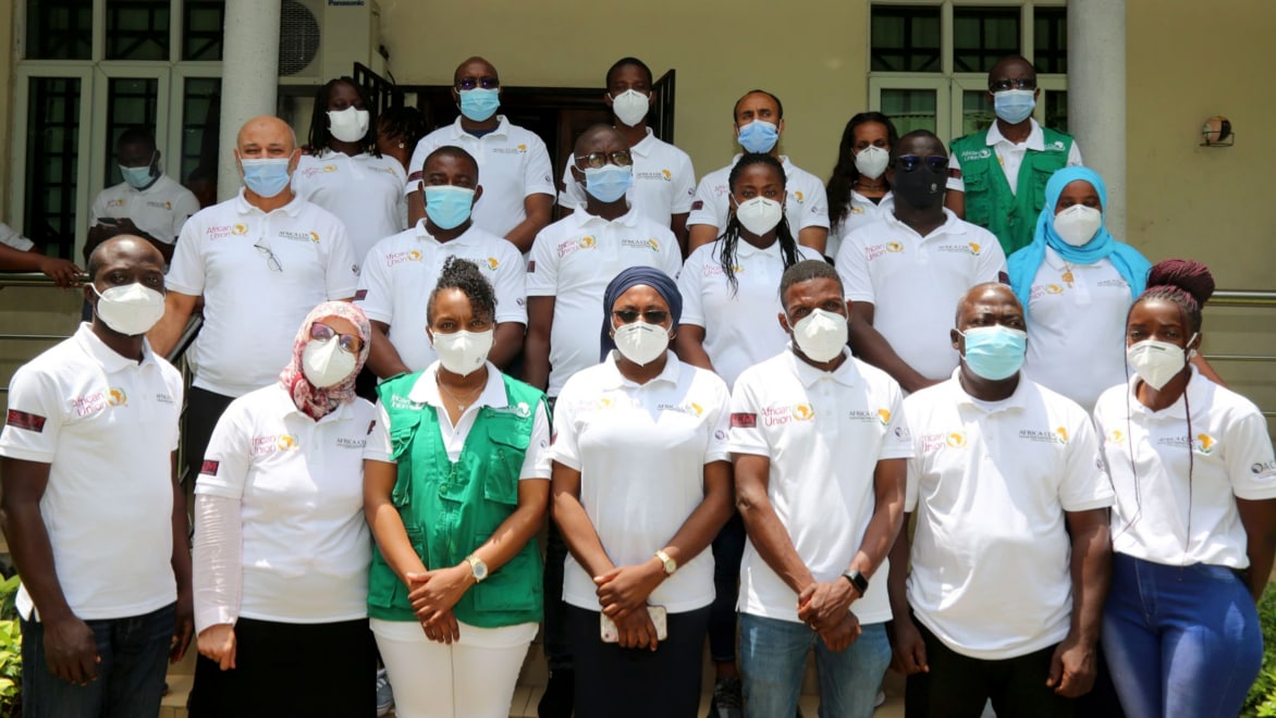 19 individuals, most of whom are wearing white "Africa CDC" polo t-shirts and white face coverings, pose for a group photo on the staircase in front of the BHI ACEGID facility.