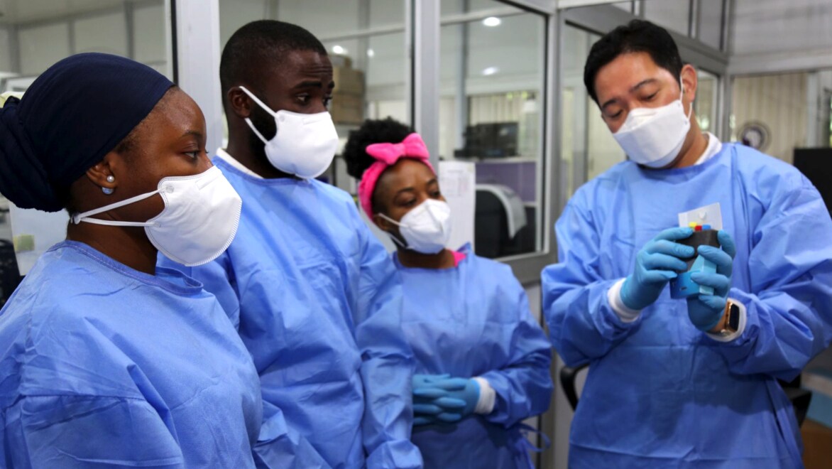 Four individuals wearing blue medical smocks and white face coverings standing in the BHI ACEGID facility. The three individuals on the left appear to be in training, looking at an object in the hands of the individual on the right, who appears to be an instructor.