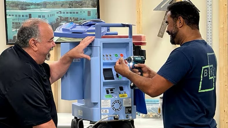 Side view of two BHI employees (men) wearing dark t-shirts, smiling, working on a blue hospital machine.