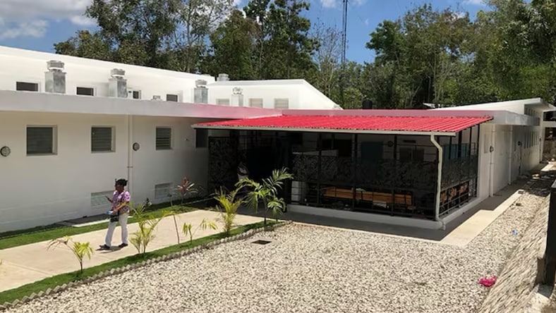Side view of a single-story BHI building surrounded by gravel and trees. A woman holding a cell phone in her right hand walking on the sidewalk next to the exterior wall.