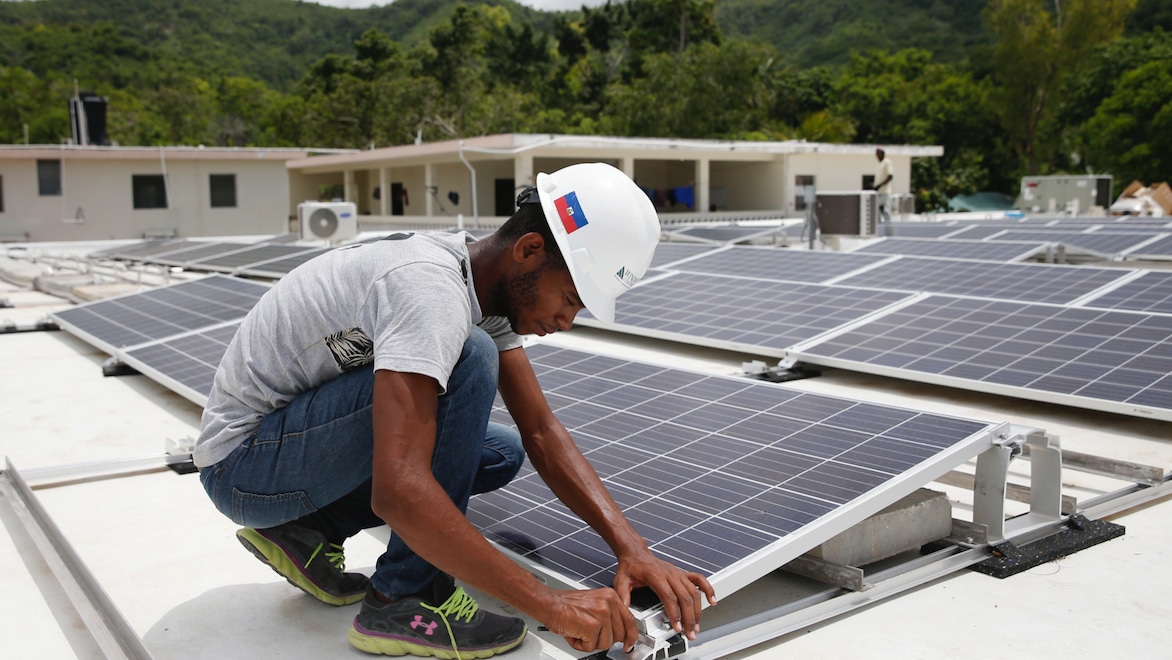 Man wearing blue jeans, sneakers, a t-shirt, and white hardhat kneeling down to work on panel of rooftop solar array, with lush green trees and mountains in the background.
