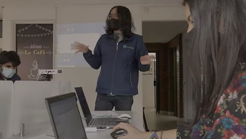 A Build Change employee standing in a classroom with a black face covering, gesturing with both her hands while providing guidance to students. Foreground: a woman student wearing a red face covering a floral shirt sitting at a computer desk. Another male student sitting to the right of the instructor, wearing a light blue face covering.