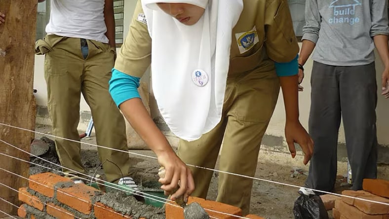 A woman wearing a white head covering bending forward toward the camera, using a masonry trowel to spread wet concrete over another layer of brick on a short brick wall, under construction at a Build Change construction site. Three male individuals, one wearing a Build Change long-sleeve t-shirt, stand in the background.