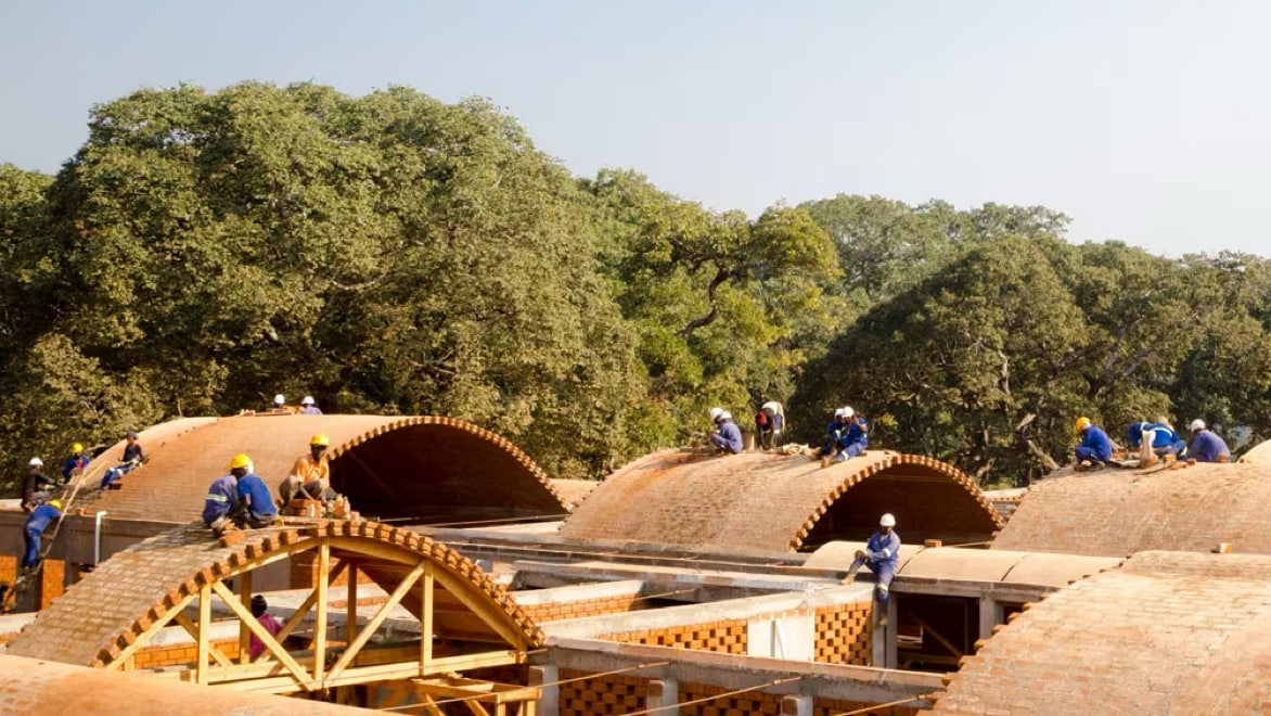 Landscape view of BuildX project with five arched brick rooftops and wooden framing. Roughly a dozen workers wearing hard hats are working atop the roofs, some sitting and some standing.