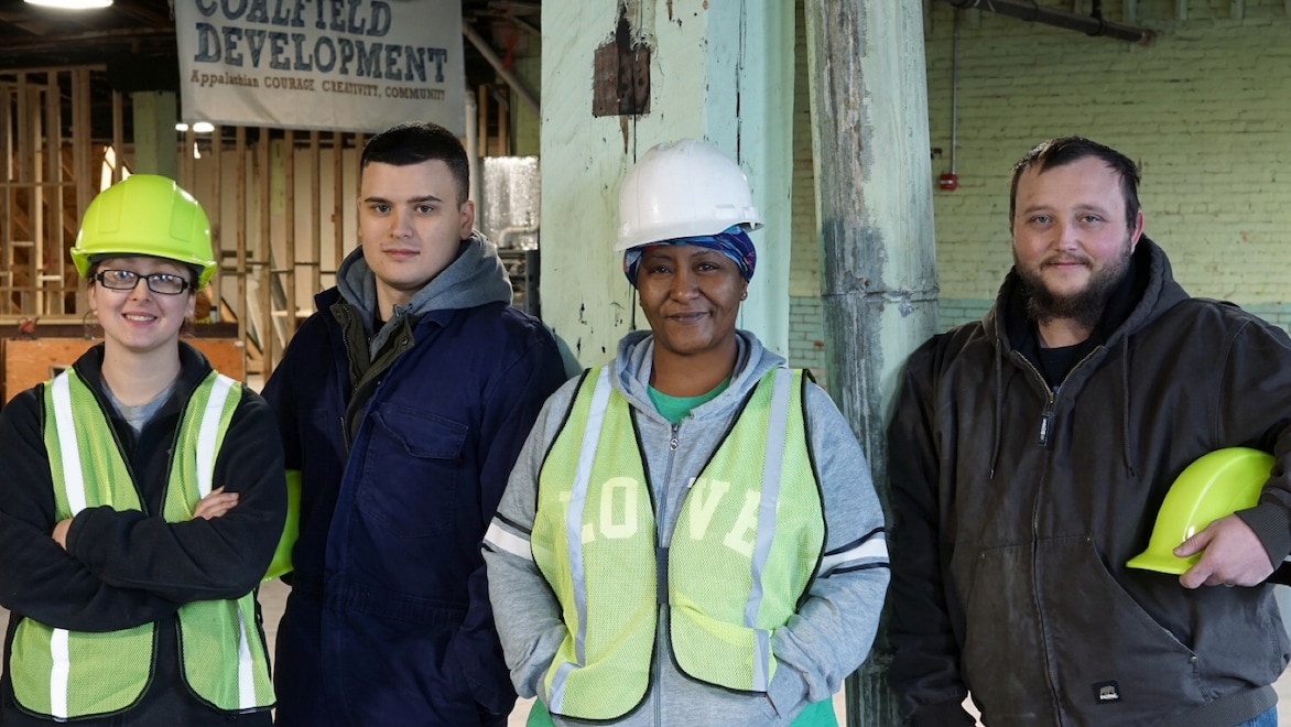 Four individuals, two women and two men, pose for a group photo wearing high-viz vests and hard hats, smiling, standing in a Coalfield Development project area (indoors).