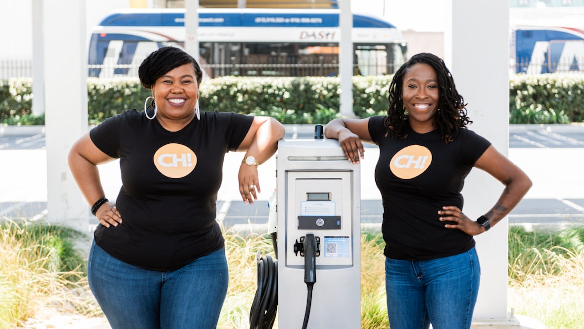 CH! Co-founders Evette Ellis (left), Chief Workforce Officer, and Kameale Terry (right), CEO, standing side-by-side, leaning against an EV charging station on a sunny day, smiling, wearing black CH!-branded t-shirts and blue jeans.