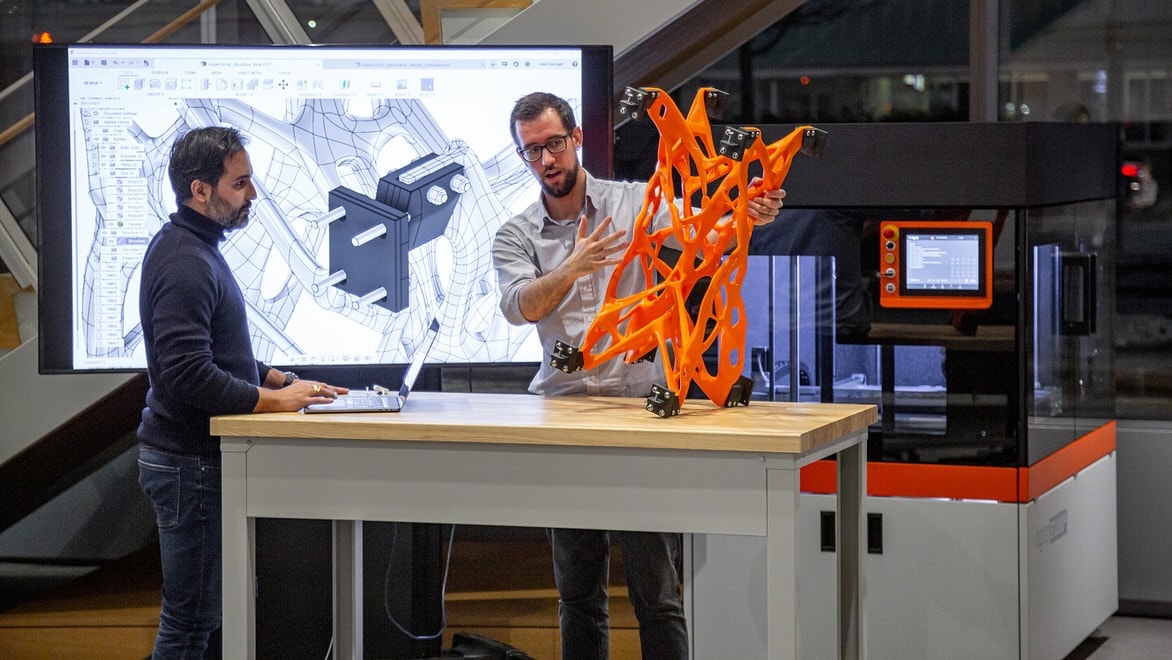 Two men examine a 3D-printed structural element while standing in front of a large screen displaying 3D modelling software.