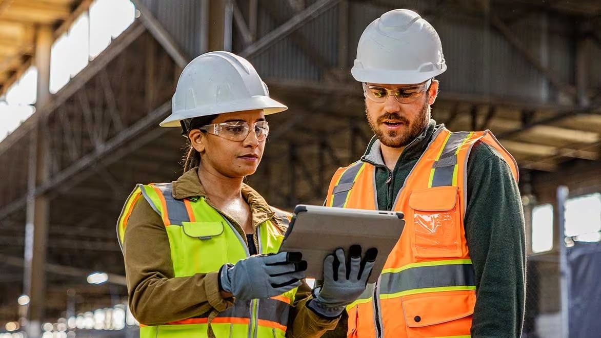 Two engineers in hard hats consult a tablet on a construction job site.