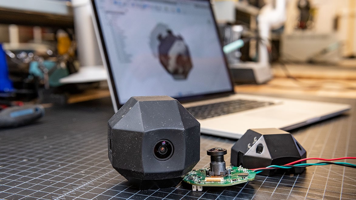 A workspace with a geometric camera device, its components, and a laptop displaying its 3D model.
