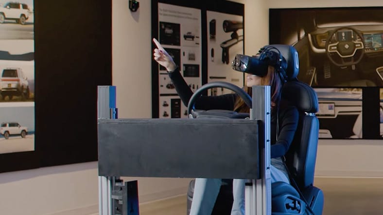 A person wearing a virtual reality headset sits in a driving simulator setup, holding a steering wheel and pointing forward, surrounded by tech displays.