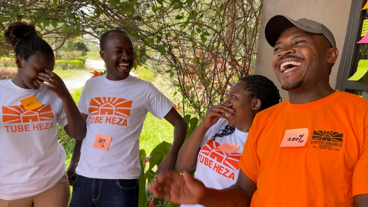 Four EarthEnable employees smiling and laughing, wearing orange and white EarthEnable t-shirts, standing on a porch of an EarthEnable project site on a sunny day. Foliage and a driveway in the background.