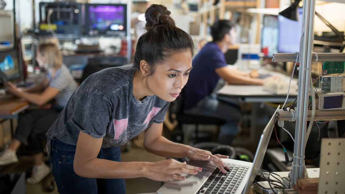 A woman wearing a grey t-shirt and jeans standing in a computer lab, leaning forward to work on a laptop. Two other people appear in the blurred background, surrounded by more computers and other tech equipment.