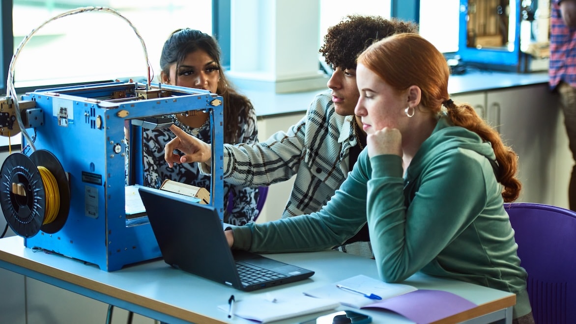 Three college students work together in a classroom in front of a laptop and a 3D printer.