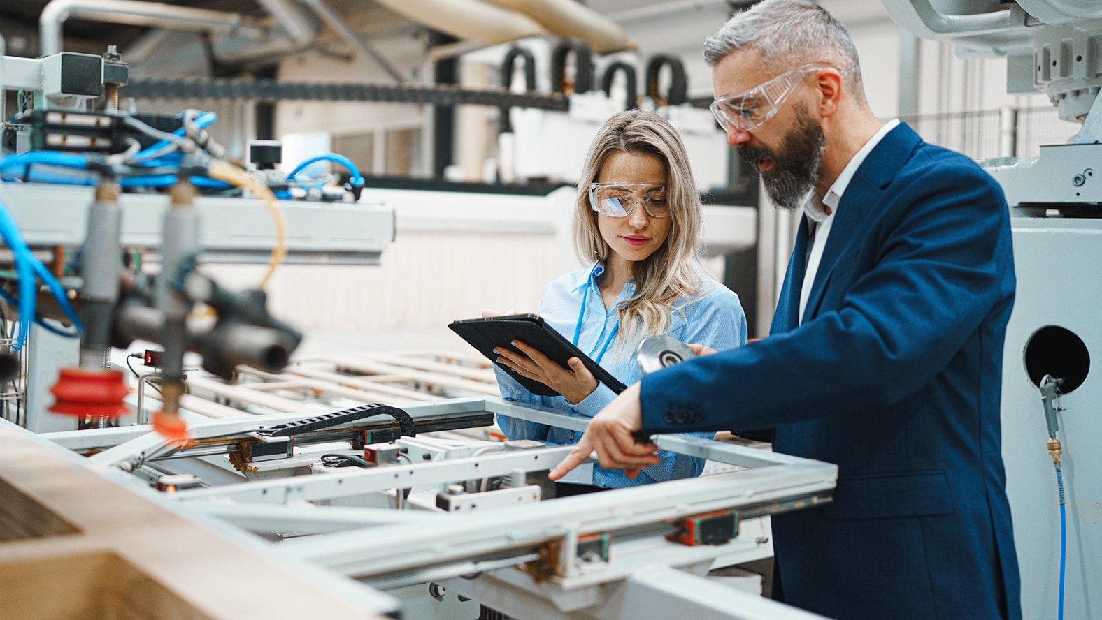 Man in suit and woman in lab coat wearing safety glasses inspecting machinery with tablet in industrial setting.