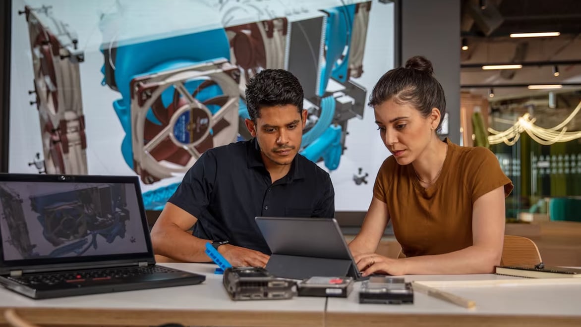 Two people working on CAD designs using a laptop and tablet in a modern workspace.