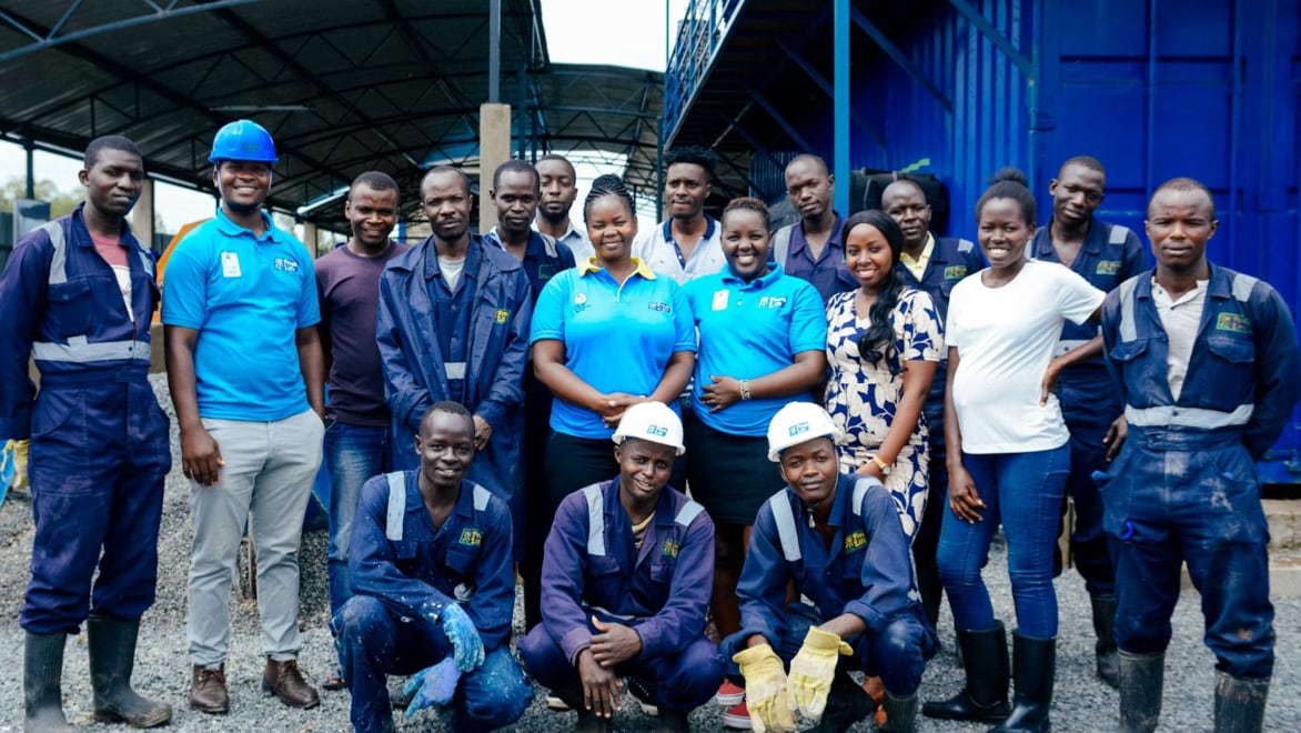 Group photo of 18 Fresh Life employees, most of whom are wearing blue Fresh Life-branded shirts or jumpsuits, standing in front of a large blue building.