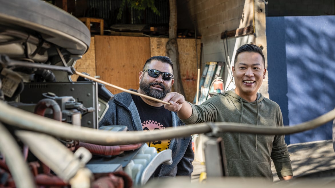 Jason Wang, FreeWorld CEO and Founder, holds a metal rod while working on a truck engine, smiling. A FreeWorld trainee stands to his right, wearing sunglasses, a black t-shirt and grey hoodie, also smiling.