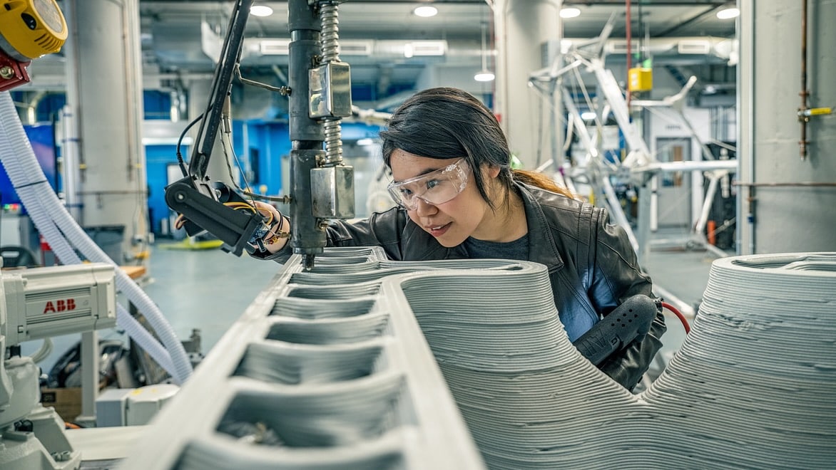 A woman works with an industrial 3D printer with a large extrusion arm layering material.