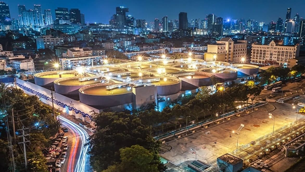 A nighttime aerial view shows an urban wastewater treatment plant illuminated with bright lights and surrounded by buildings, greenery, and roads.