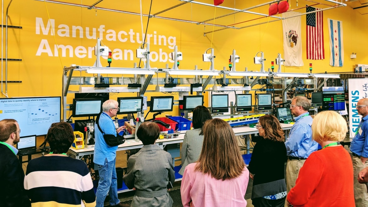 A group of Hope Street Group trainees facing away from the camera toward an instructor—an older man with grey hair, glasses, and a black sling around his right arm—who is pointing at a row of machines and computer monitors under a large "Manufacturing American Jobs" wall sign.