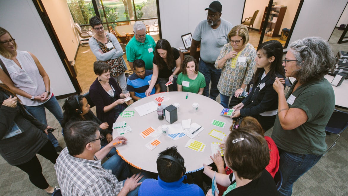 Seventeen individuals of varying ages and diverse ethnicities sitting and standing around a round table with sticky notes and writing implements during a workshop hosted at The Industrial Commons.