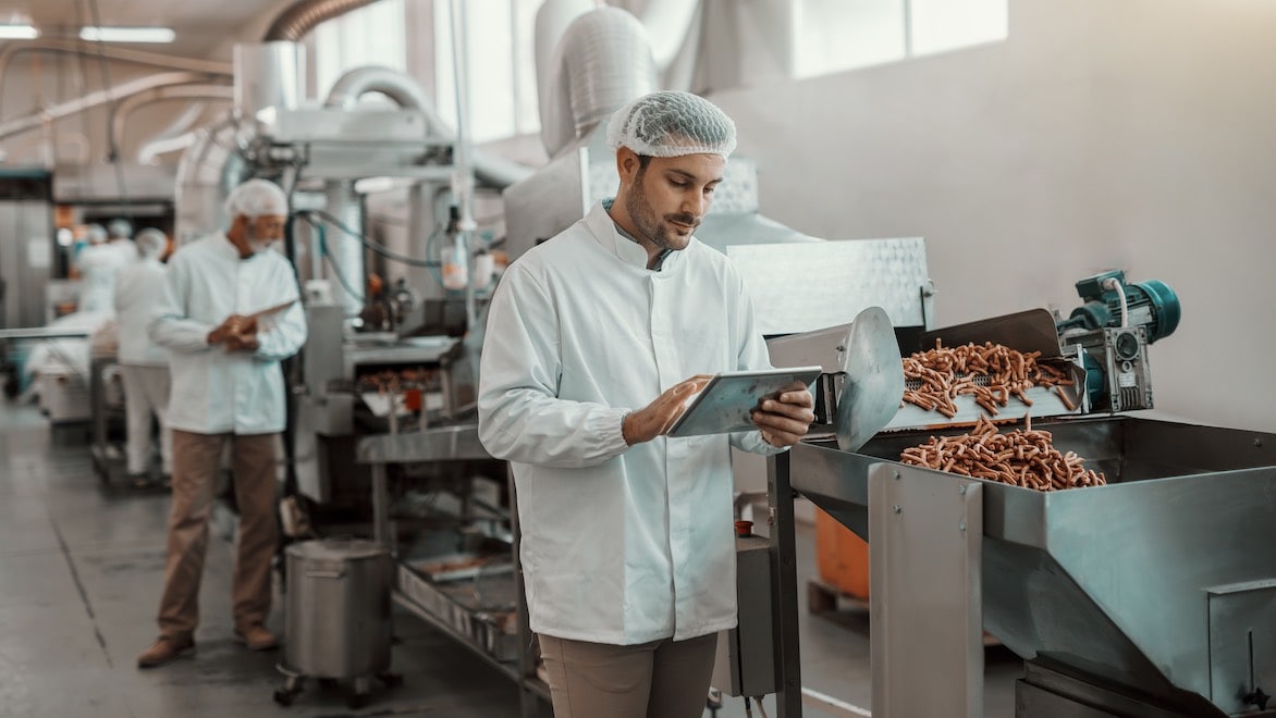 Man in white jacket and hairnet holding tablet while inspecting food in a food processing plant.