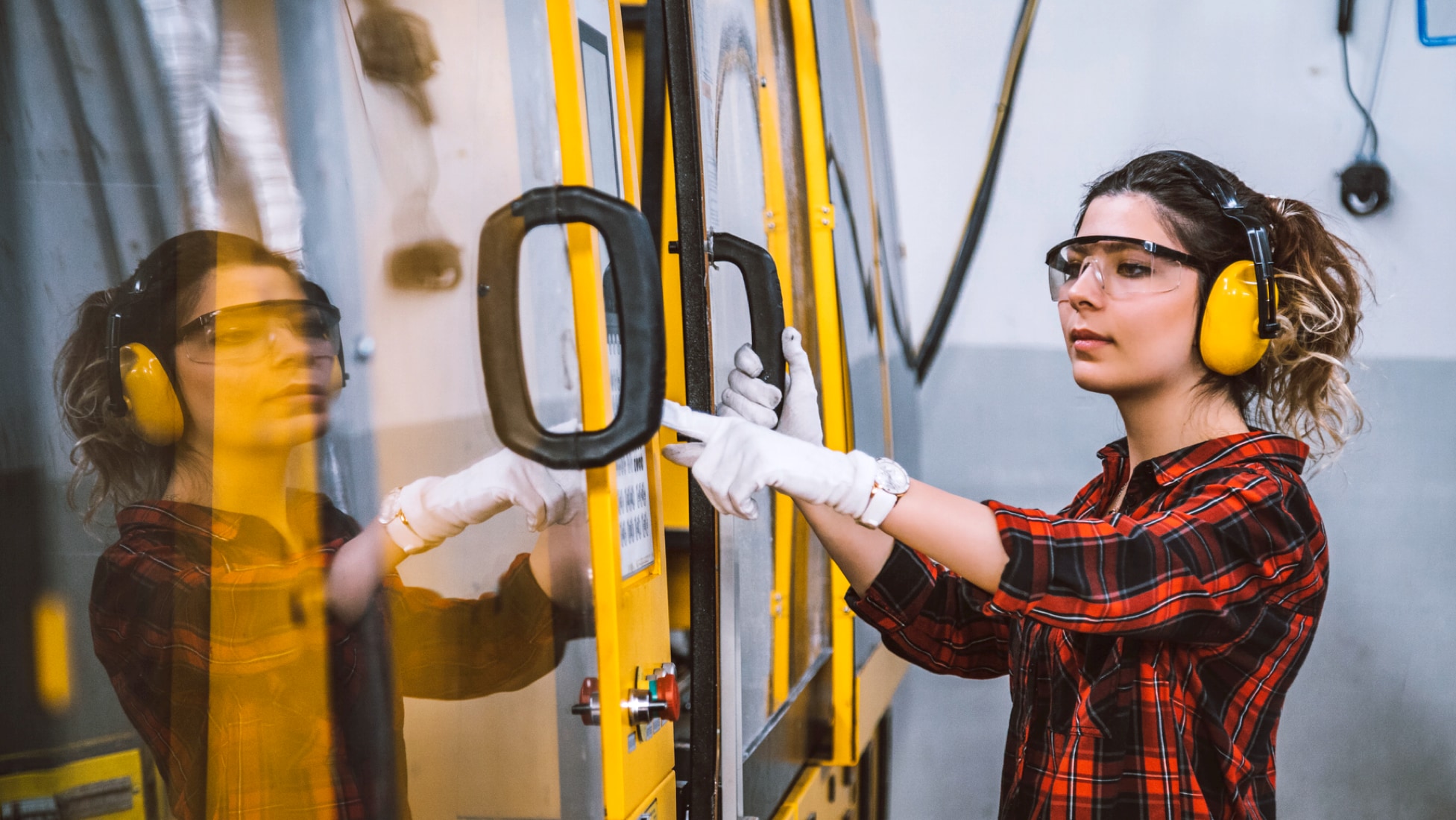 Side view of a woman wearing safety glasses, hearing protection, and white gloves, standing next to a CNC machine, holding onto a black door panel handle with her right hand and pressing buttons on a control panel with her left hand, wearing a red, blue, and black flannel. Her brown and blonde hair is held back in a ponytail. Her reflection is showing in the glass door of the machine (left side of photo). The room is fluorescent-lit.