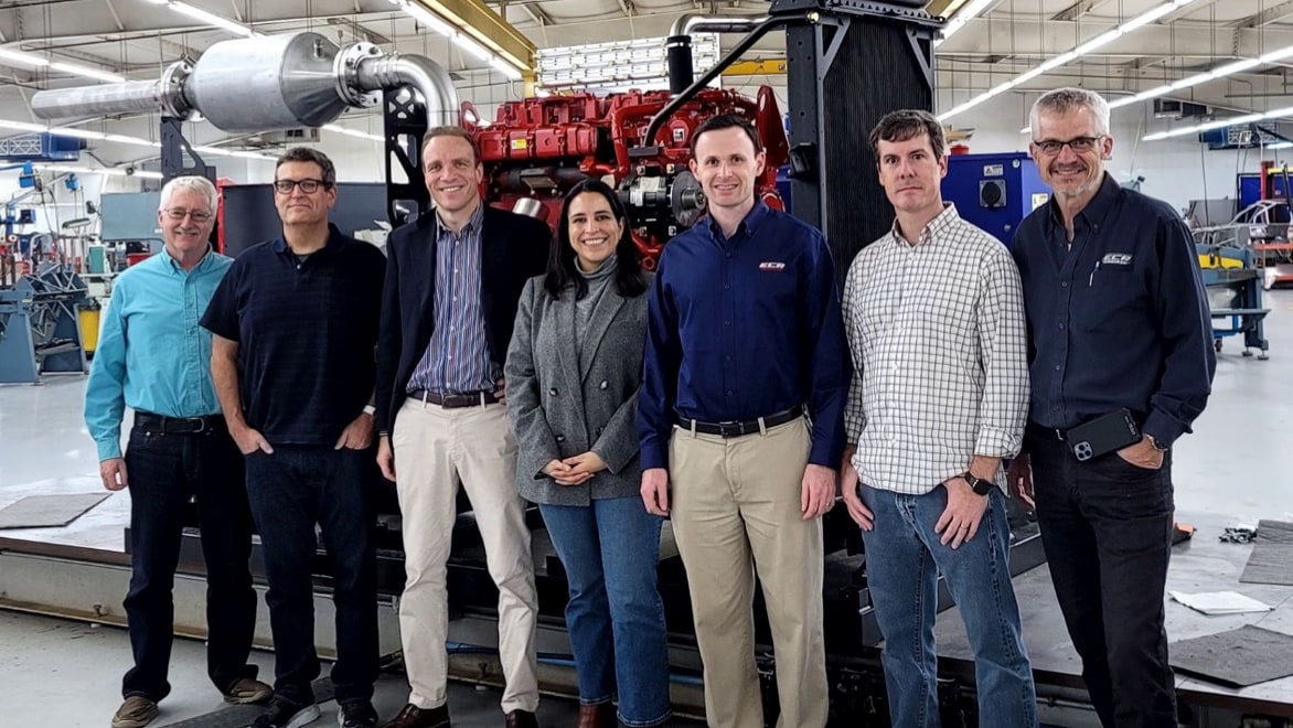 The M2X Energy team standing in warehouse facility, posing for a group photo in front of an M2X methane-to-methanol conversion unit, a large truck-sized contraption that looks like a large energy generator. From left to right: From left to right: Anthony Dean, PhD, COO; Josh Browne, PhD, CTO; Massimiliano Pieri, CEO; Diana Alcala, VP Business Development; Kyle Merical, Principal Engineer; Paul Yelvington, PhD, Chief Science Officer; Andrew Randolph, PhD, VP Engineering.
