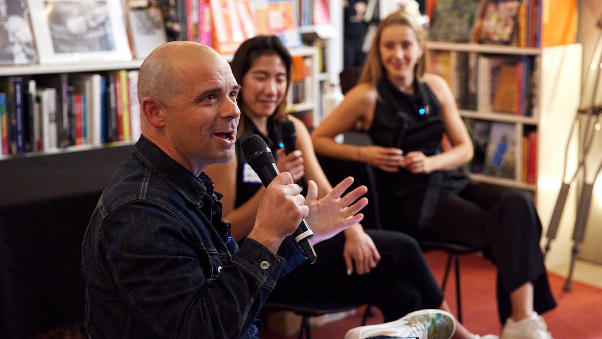 Three individuals sitting in chairs at a NYC Climate Week 2023 event with MCJ Collective on October 4, 2023. Cody Simms, an MCJ Collective Partner, sits in the foreground, in focus, wearing a denim jacket and speaking into a microphone, gesturing with his hands; to his left sit Sophie Purdom of Planeteer and Kim Zou of CTVC. The individuals are sitting in a book-lined event space with natural and artificial light.