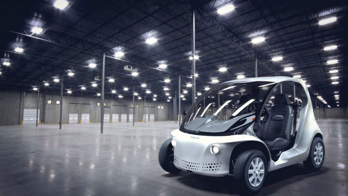 A compact, futuristic electric vehicle (EV) inside a spacious industrial warehouse.