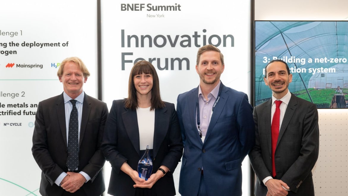 BloombergNEF CEO Jon Moore (left) and Global Head of Innovation, Client Relations and Partnerships Benjamin Kafri (right) presenting Nth Cycle CEO Megan O’Connor (middle-left) and CFO Coleman Adams (middle-right) with a 2023 Pioneers Award at the BNEF Summit, April 24, 2023. All four individuals are smiling, wearing blue or black blazers, standing in front of a pull-up banner that reads, "BNEF Summit: New York" (top) and "Innovation Forum" (larger font, below).. Megan is holding the BNEF award with both her hands supporting the base of the blue/clear glass award. 