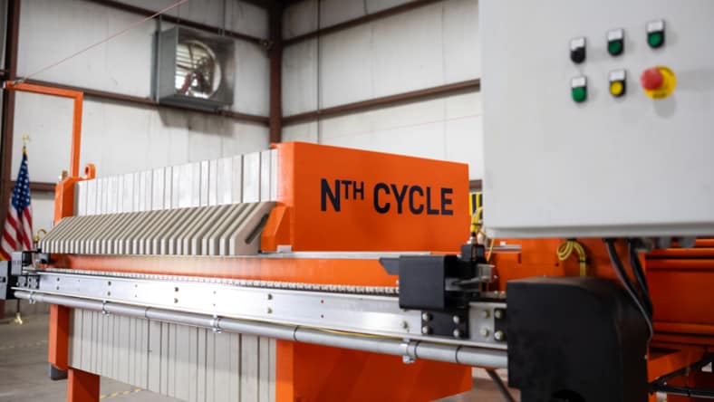 Side view of Nth Cycle's OYSTER electro-extraction processor in Nth Cycle facility. Colored buttons on a metal panel at the top-right corner. Focal point: Nth Cycle logo (black text) on side of OYSTER unit (painted orange). No ceiling visible. Free-standing American flag in the background to the left of the OYSTER.