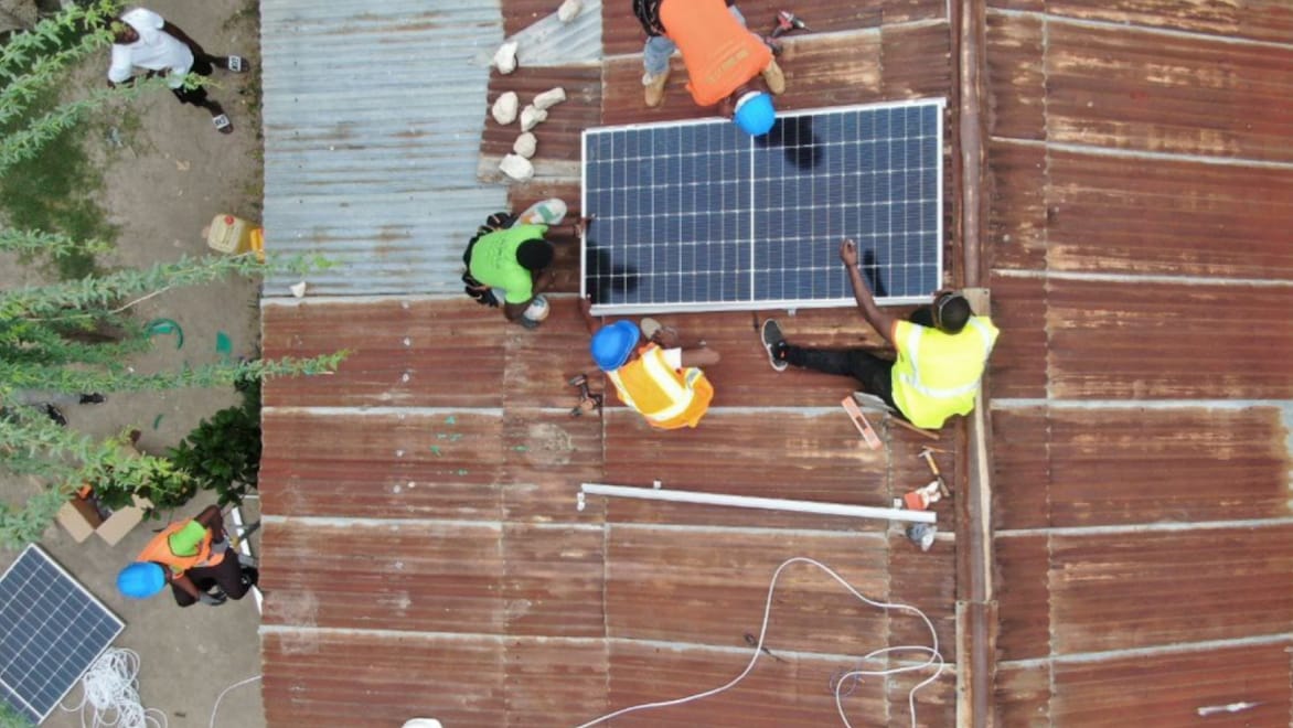 Aerial view of four Okra Solar workers installing a large solar panel on a rusty corrugated metal roof. Two individuals standing on the ground and watching the installation.