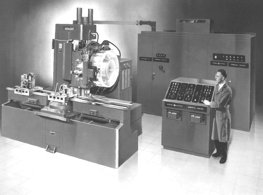 A black and white photograph from the mid 20th century shows inventor John T. Parsons standing next to a large prototype CNC machine consisting of several control cabinets and a milling apparatus.