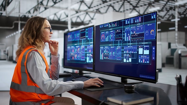 Person wearing an orange safety vest and glasses sitting at a desk with two large monitors displaying complex technical data in an industrial setting