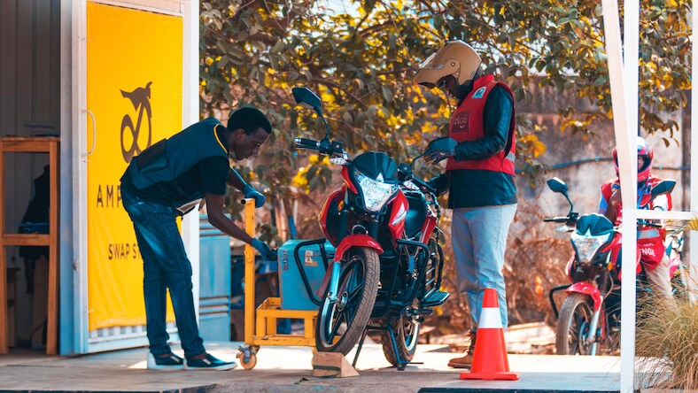 Two men working on a motorcycle under an awning.