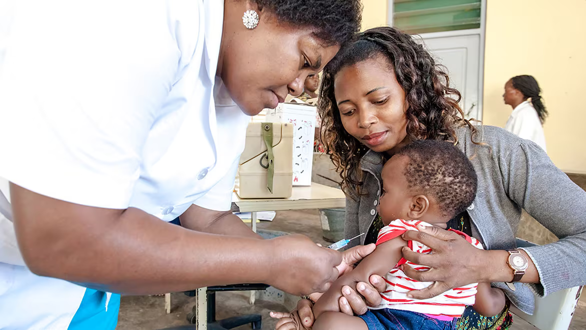 A woman healthcare provider holding a syringe, about to administer a vaccine to an infant who is being health by a woman guardian.