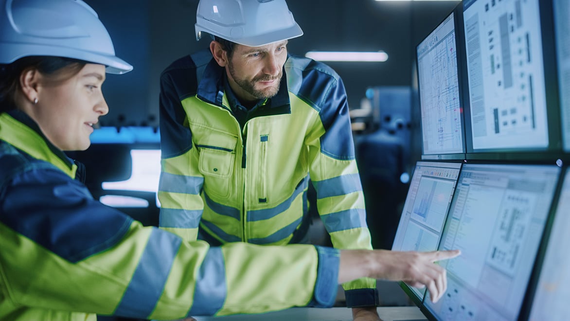 Two manufacturing facility managers wearing hard hats inspect data on control screens.