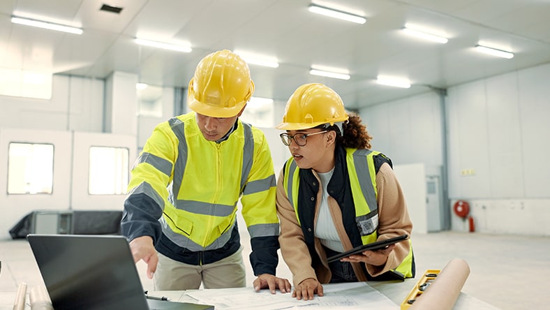 Two construction workers wearing yellow hard hats and reflective vests reviewing plans on a table in a bright indoor space