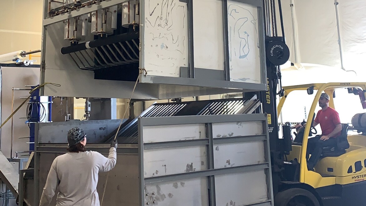 Two Rebound Technologies employees work together to maneuver to dumpster-sized components of the IcePoint machine. One employee is using a forklift to place the top component onto the bottom while the other employee helps guide the placement using a rope.