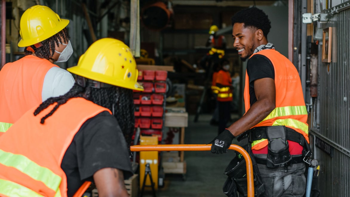 Three young men—two wearing yellow hard hats facing away from the camera, one facing toward the camera, smiling and wearing work gloves—tag-team moving an orange cart through a warehouse. All are wearing orange and yellow hi-viz safety vests.