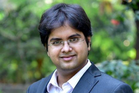 Headshot of Dr. Vaibhav Tidke, Co-Founder & CEO, S4S Technologies. Man with black hair and rimless glasses, looking at the camera and smiling, wearing a white collared shirt and a black blazer, standing outside on a sunny day, with trees in the blurred foliage in the background.
