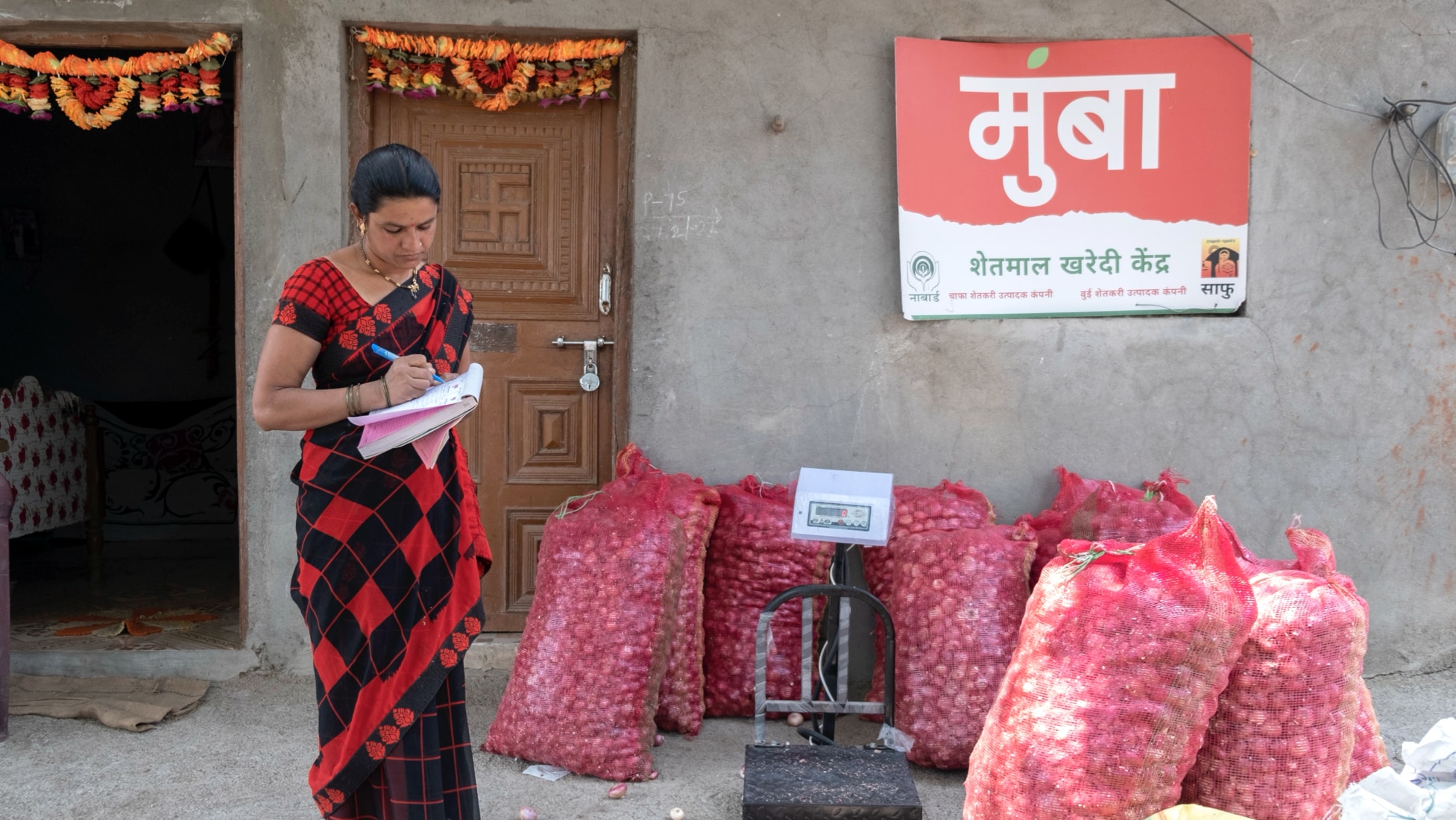 A woman wearing a red and black saree (traditional Indian dress) standing in front of a building with a red door, holding a notepad in her left hand and a blue pen in her right hand, looking down at the notepad as she writes. To her left are ~10 large sacks of produce leaned against each other in front of the building, next to a scale.