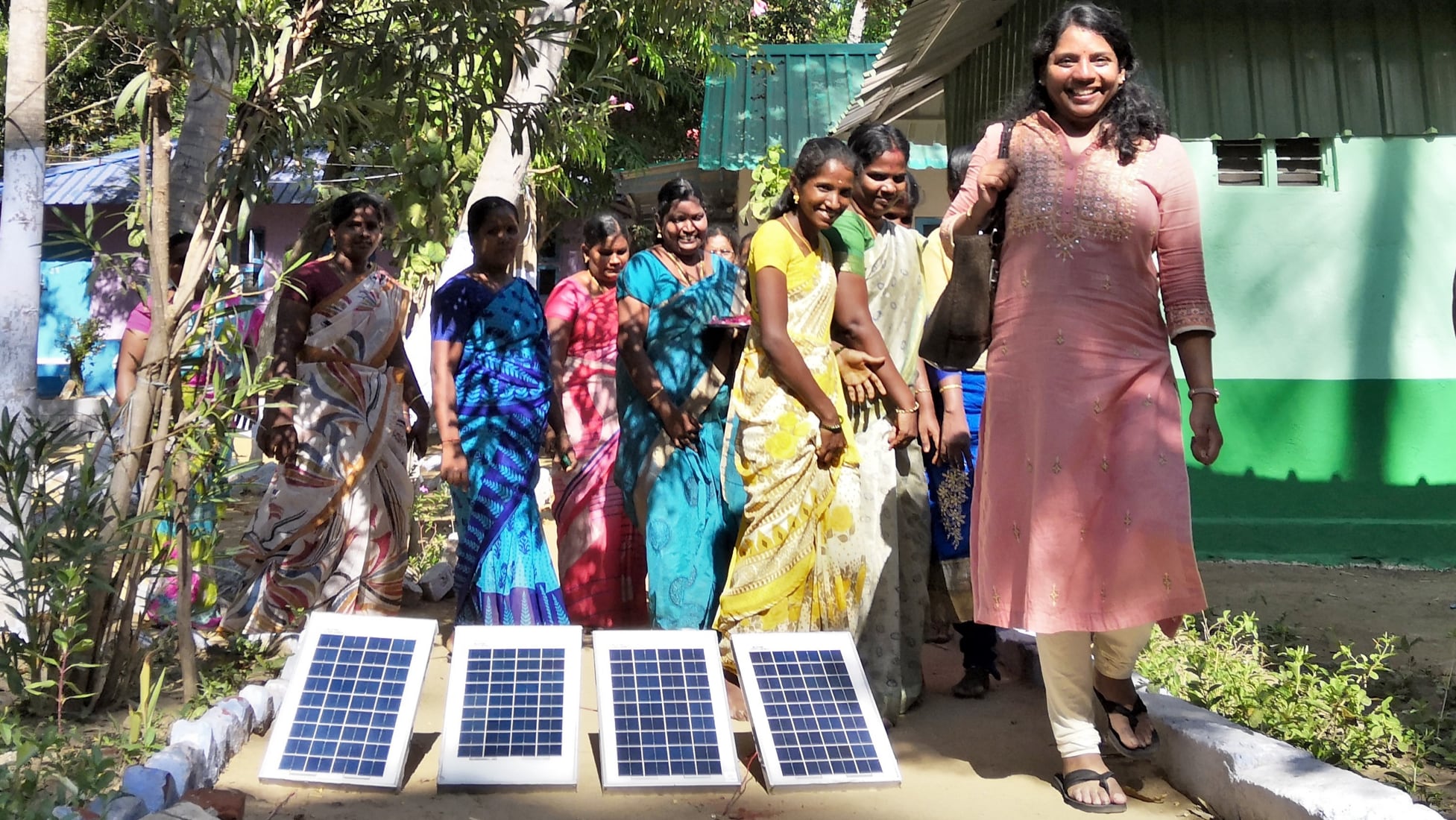 Eight Indian women smiling, wearing brightly colored dresses, standing behind four small solar panels in a row on the ground. Sunlight shining through trees onto the women and the solar panels.