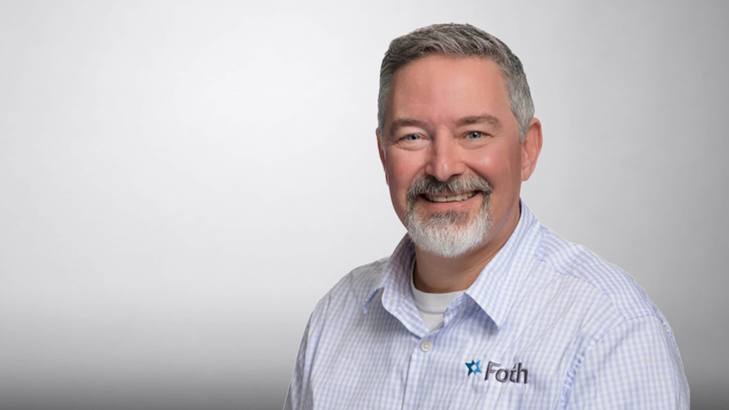 Smiling man with gray hair and beard wearing a white shirt with 'Foth' logo on chest.