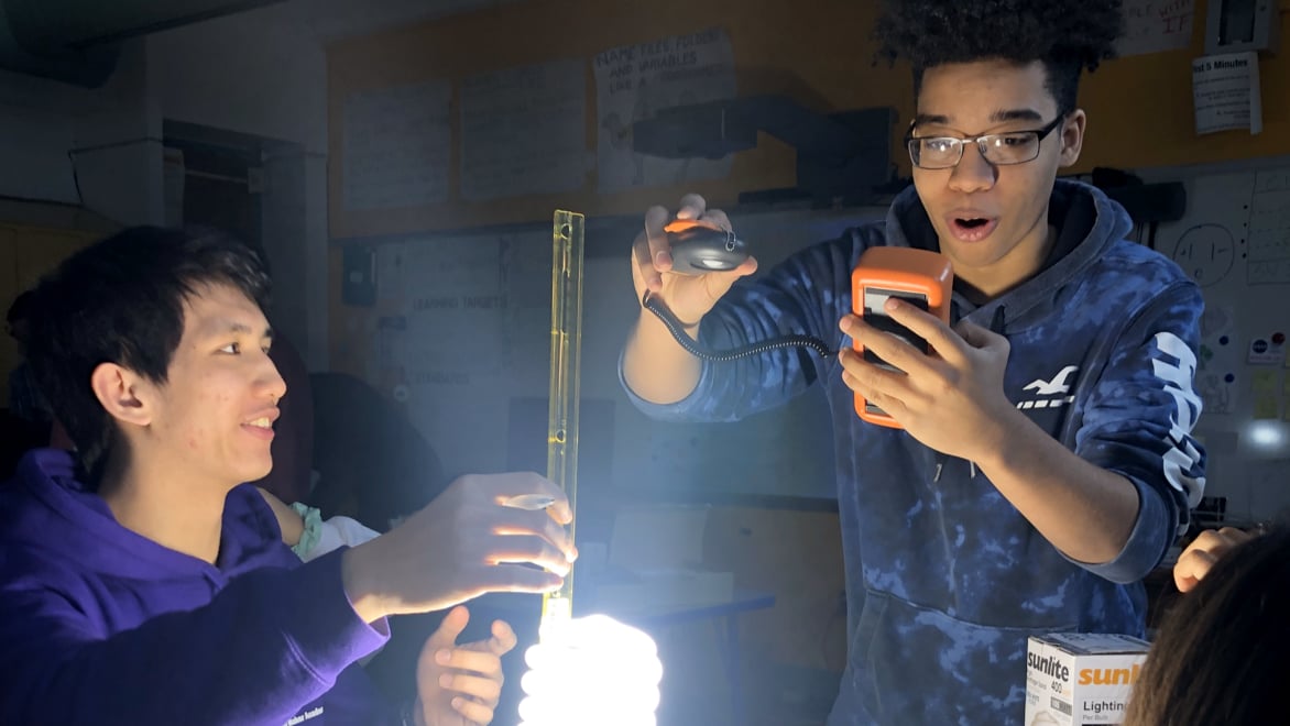 Two male students conducting an experiment with a light bulb and light meter in a darkened classroom. The student on the right is looking at a light meter with an expression of awe; the student on the left is holding a ruler, measuring the distance of the meter from the light bulb, smiling and looking at the other student.