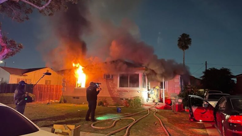 A house engulfed in flames with firefighters and media personnel on-site.
